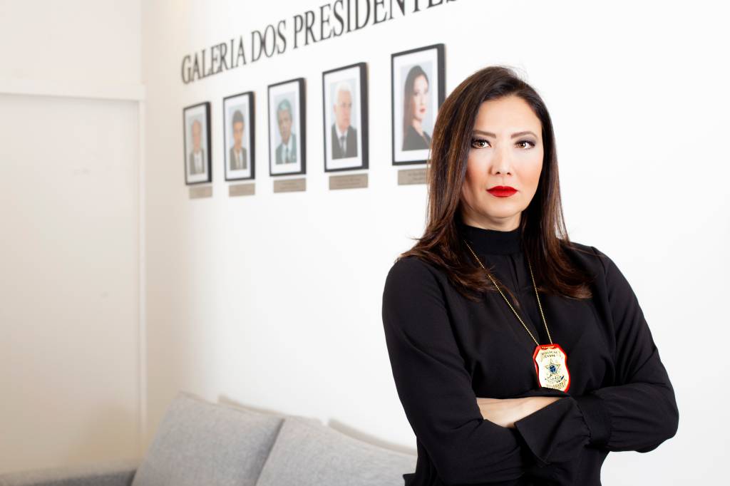 Raquel Gallinati, presidente do Sindpesp, posa ao lado dos retratos dos presidentes anteriores vestindo um suéter preto e batom vermelho.