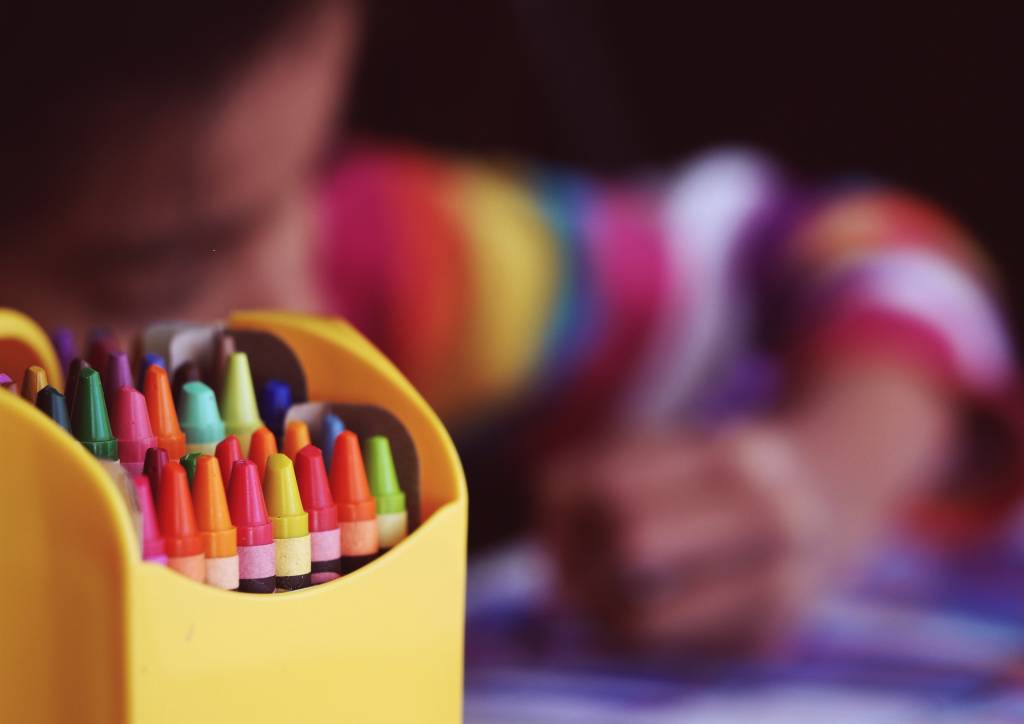 Imagem mostra caixa de lápis de cor em primeiro plano e criança pintando desenho com lápis de cor ao fundo