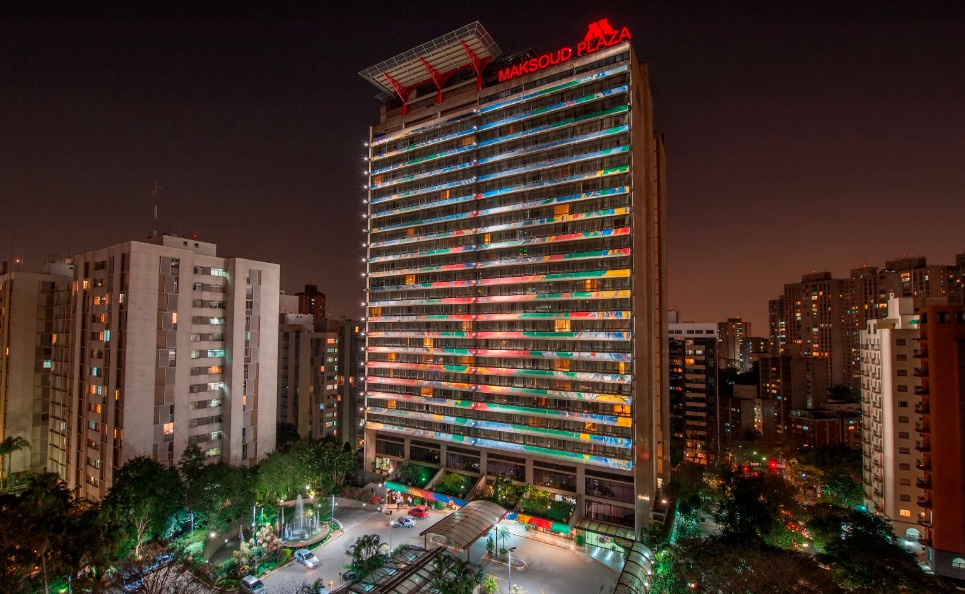 Foto aérea exibe fachada inteira do Hotel Maksoud Plaza com pintura colorida do prédio e espaço do estacionamento.