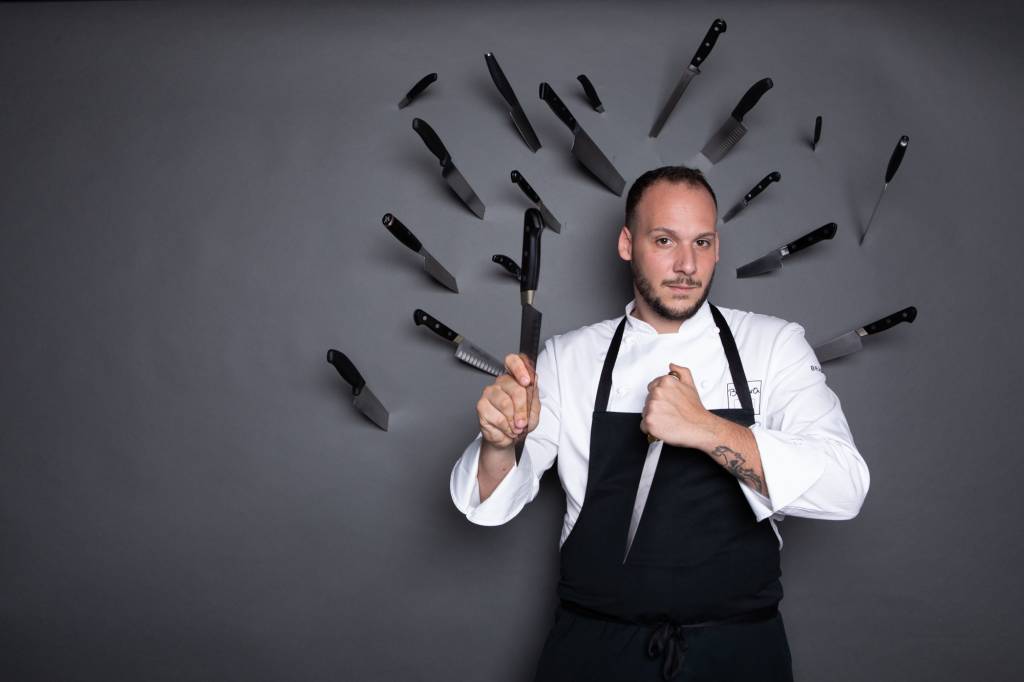 Chef de avental com facas na mão e na frente de parede cheias de facas