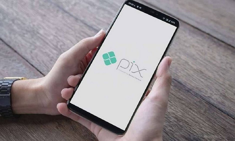 A imagem demonstra uma mão segurando um celular com a tela virada para cima em que está presente o logo do PIX.
