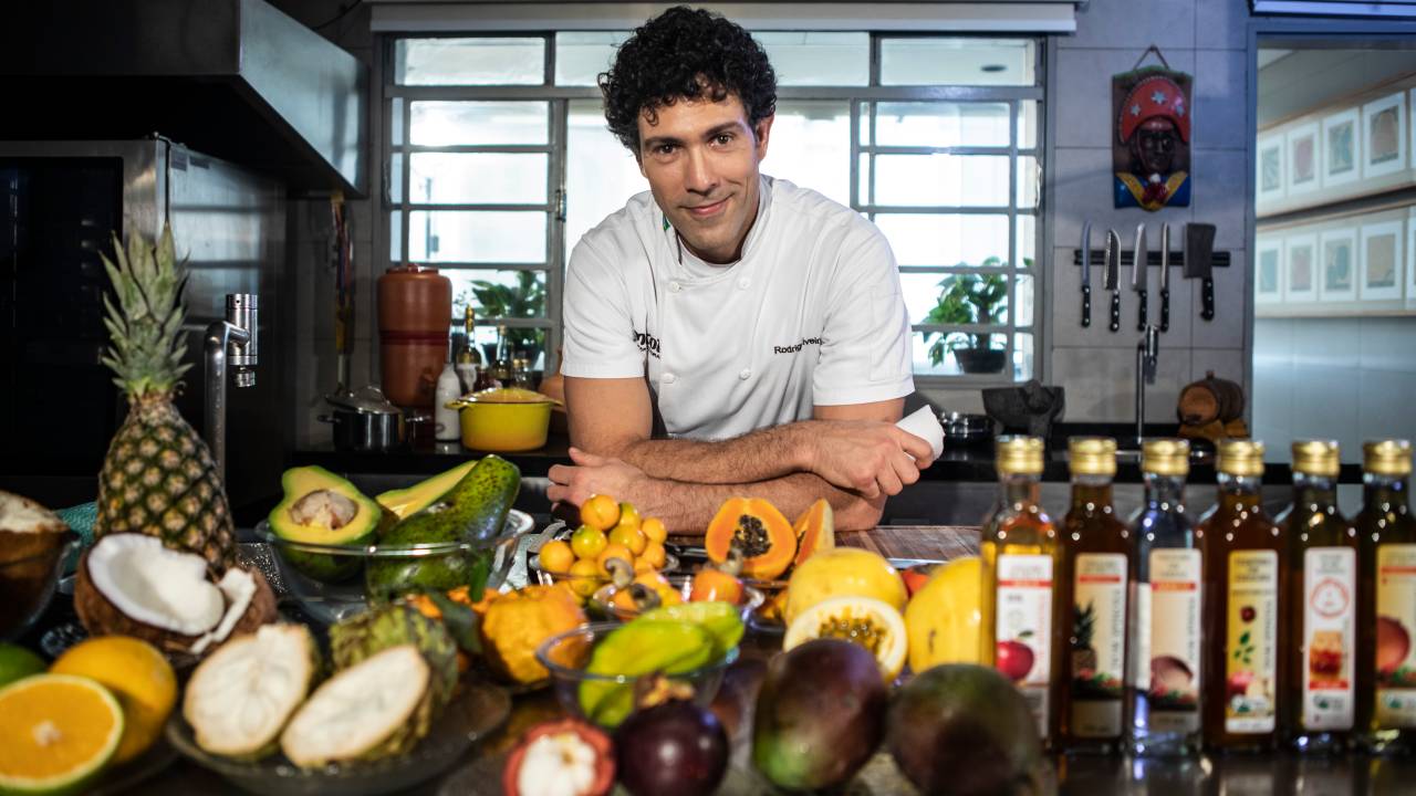 Rodrigo Oliveira, do Mocotó, posa encostado em mesa repleta de ingredientes, incluindo frutas e azeites.