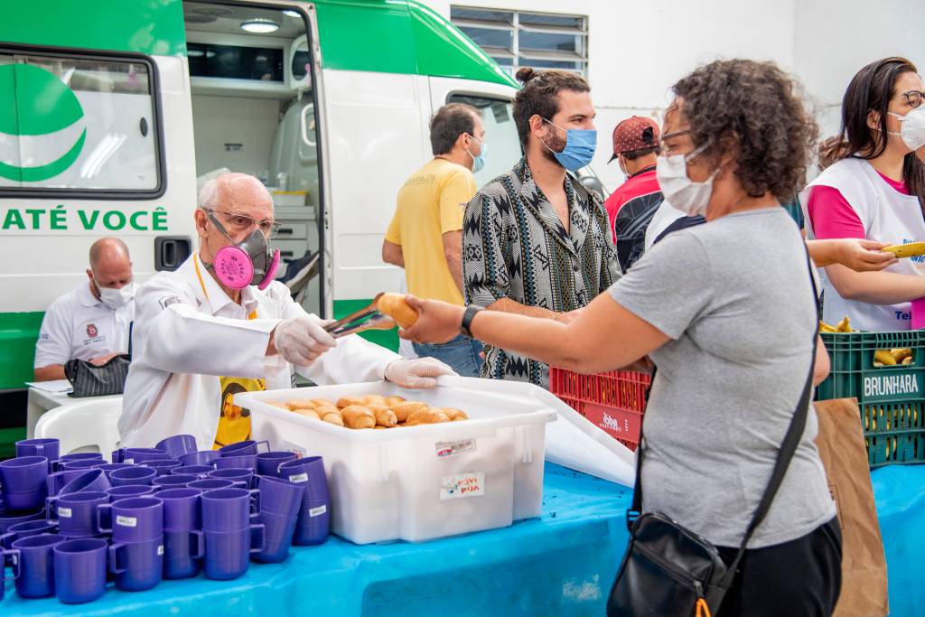 Padre Júlio Lancellotti, de máscara, distribuindo pão com pinça de uma caixa de plástico branca com pães.