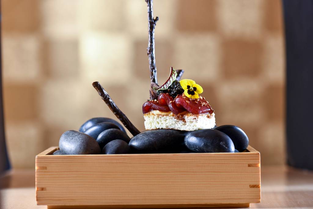 Fatias de atum marinado sobre uma torrada de brioche, maionese de peixe e caviar, em cima de pedras pretas dentro de caixa de madeira.