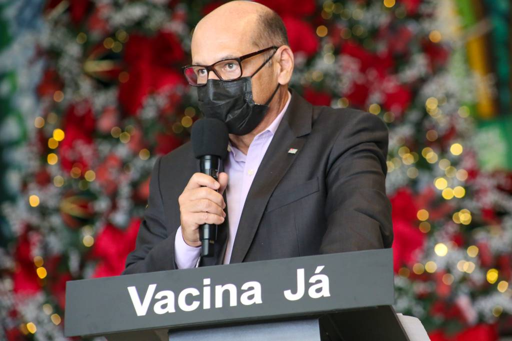 Imagem mostra Dimas Covas de terno, em púlpito com inscrição "Vacina Já". Ele usa tblazer e máscara e segura microfone