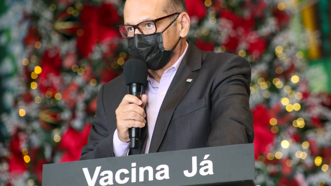 Imagem mostra Dimas Covas de terno, em púlpito com inscrição "Vacina Já". Ele usa tblazer e máscara e segura microfone