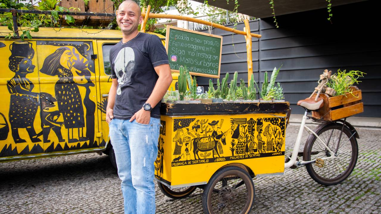 Israel Joaquim posa de pé entre sua perua e bicicleta amarelas com ilustrações pretas inspiradas em xilogravuras nordestinas. Veste calça jeans e camiseta preta estampada com uma caveira.