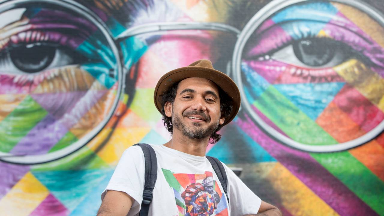 Eduardo Kobra sorri em frente a mural com rosto de John Lennon colorido.