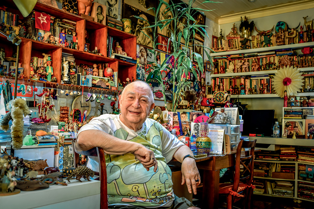 Imagem mostra Sergio Mamberti sorrindo, sentado em cadeira em quarto com dezenas de livros e brinquedos pelas paredes