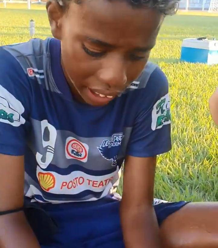 Menino de 11 anos, uniformizado, chorando em um campo de futebol