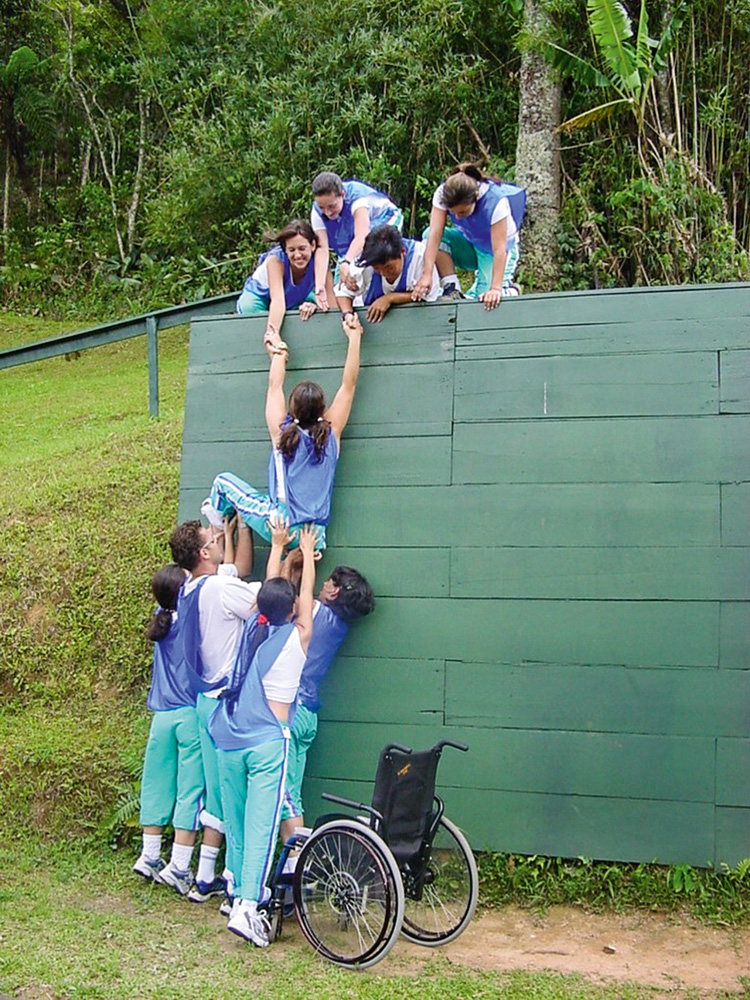 treinamento Carol Ignarra Carol treinando. Enquanto é empurrada por colegas, ela tenta alcançar a parte de cima de um muro verde, com auxílio de outras pessoas já na parte de cima