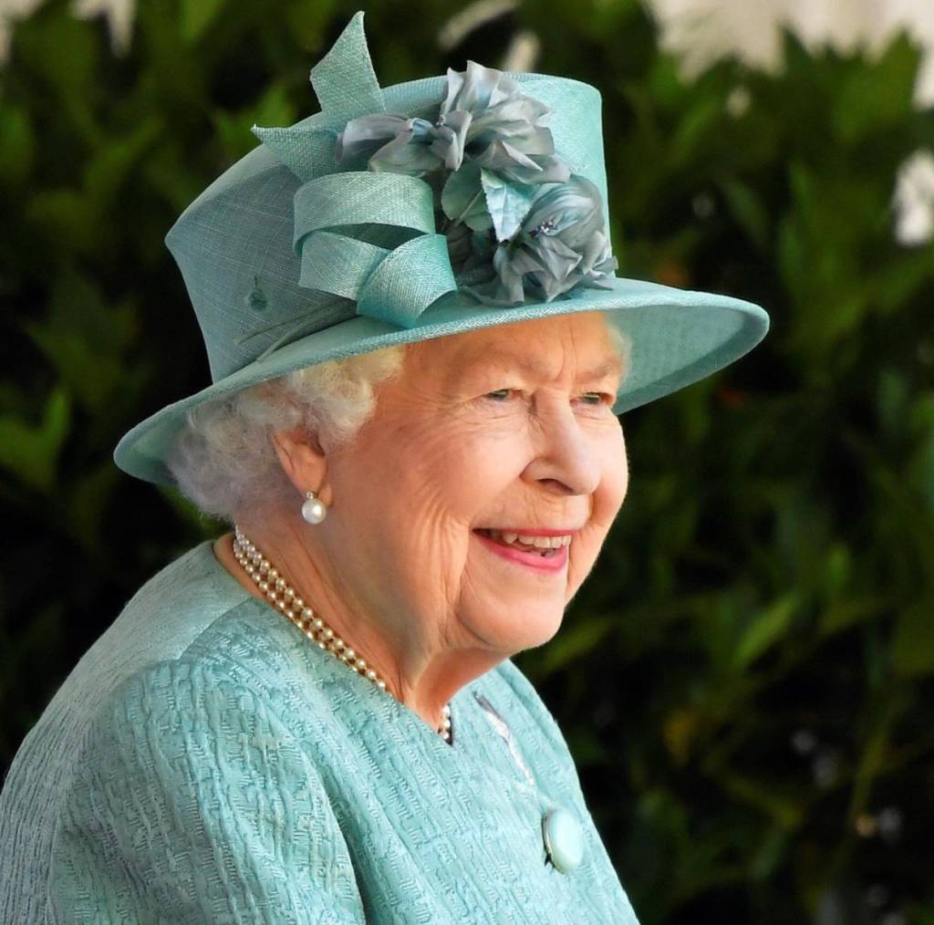 A imagem mostra Rainha Elizabeth II sorrindo. Ela usa chapéu e vestes de cor verde azulado