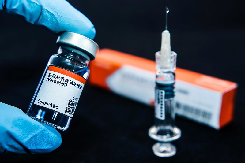 A imagem mostra uma ampola da CoronaVac sendo segurada por uma mão com luva sobre uma mesa. À frente, há uma agulha de aplicação da vacina.