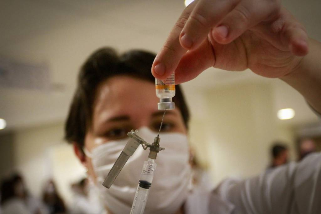 Imagem exibe mulher com frasco da vacina CoronaVac, colocando o líquido em uma seringa de vacinação