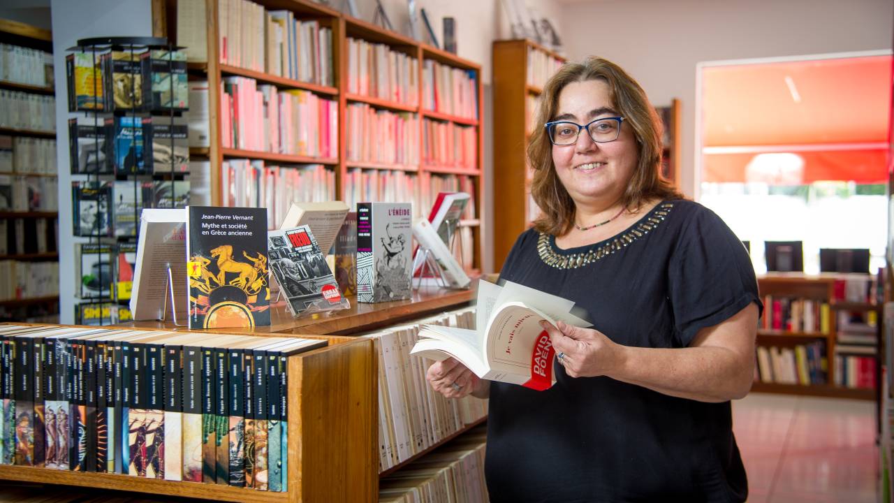 Silvia Monteil, da Livraria Francesa, posa de pé com livro aberto na mão e prateleiras repletas de livros franceses ao redor.