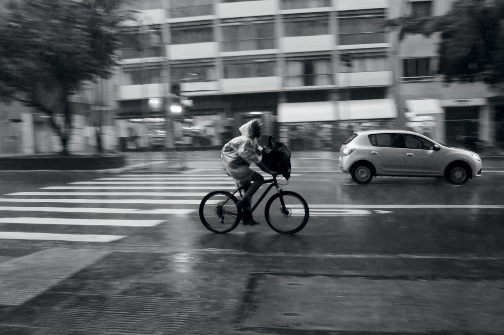 Homem andando em uma bicicleta no meio da rua durante a chuva. Preto e branco.