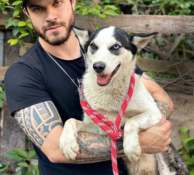 Vereador Felipe Becari veste boné, camiseta preta e segura cão de médio porte com pelos brancos e pretos na região do rosto e orelhas.