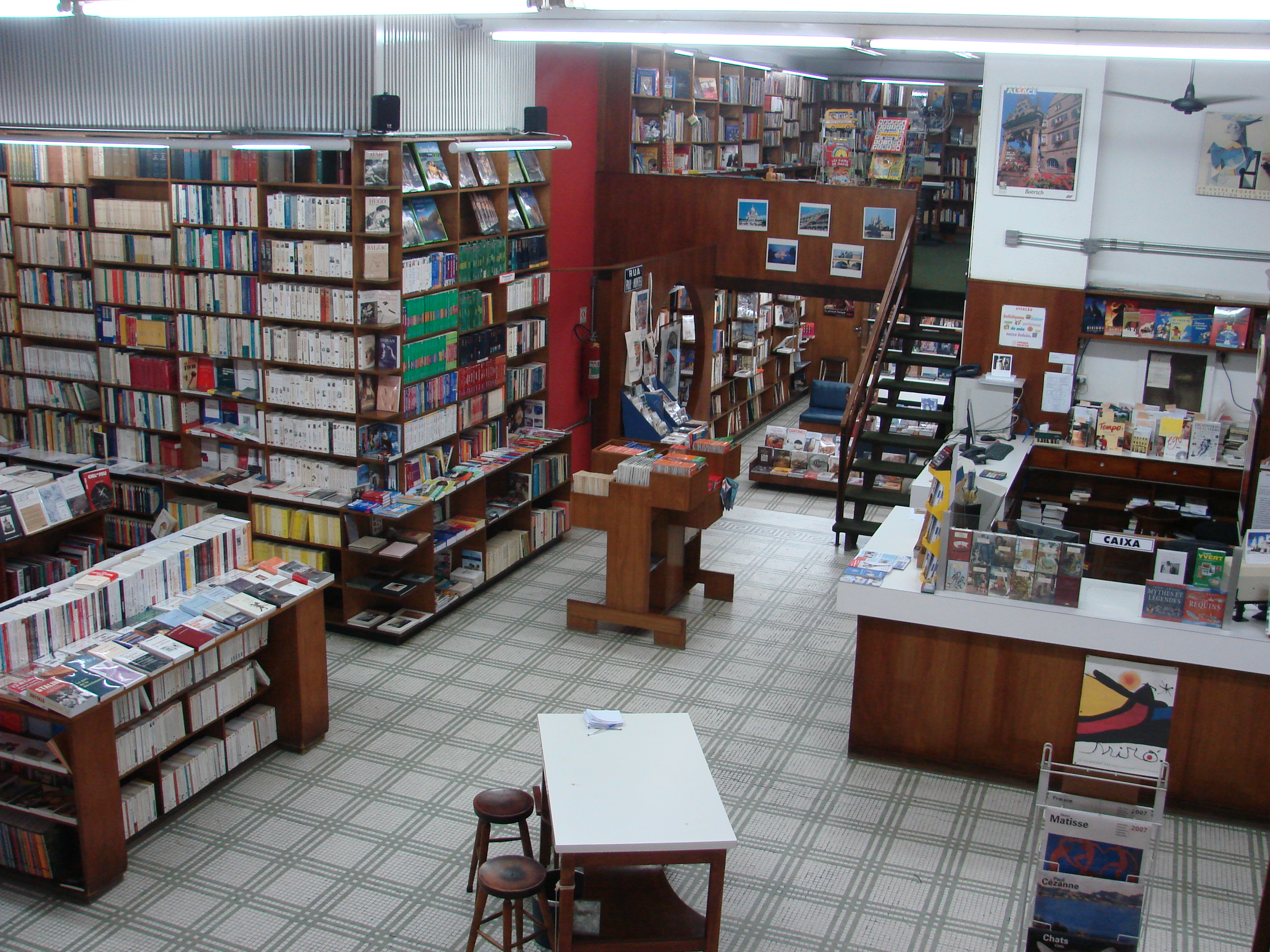 Imagem aberta exibe as prateleiras da antiga loja da Livraria Francesa no Centro, com escada no fundo e balcão à direita.