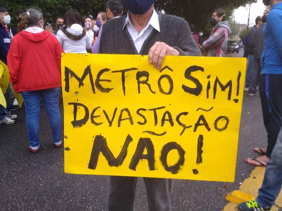 Morador do Jardim Têxtil protesta contra obra de metrô na Praça Mauro Broco com placa amarela escrito "Metrô sim! Devastação não!"