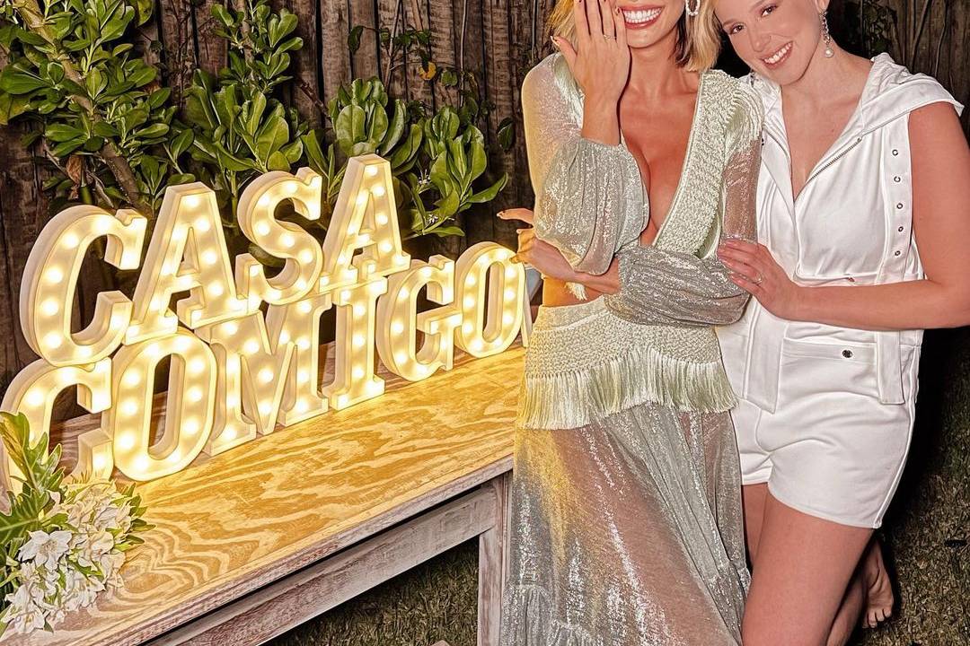 Vitória Strada e Marcella Rica posam abraçadas com roupas brancas ao lado de letreiro luminoso que diz "casa comigo".