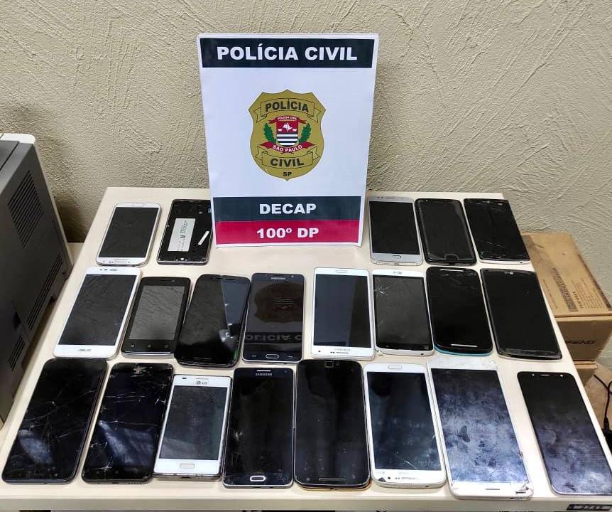 Imagem mostra celulares colocados em mesa com logo da Polícia Civil ao fundo