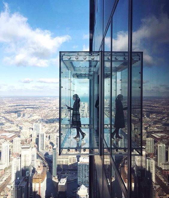 Box de vidro surge projetado para fora de um prédio com vista da cidade ao fundo. Uma mulher aparece dentro do espaço envidraçado olhando para baixo.