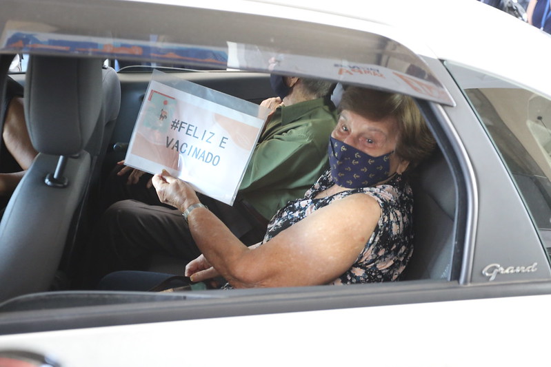 A imagem mostra uma idosa, com uma placa escrito "feliz e vacinada". Ela está dentro de um carro.