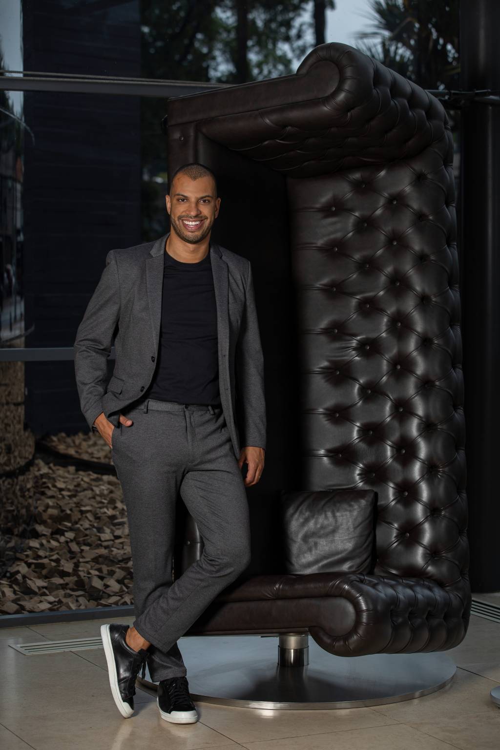 Wellington Melo, do Unique, posa de pé ao lado de sofá de couro preto erguido de pé como escultura. Veste terno cinza com camiseta preta.