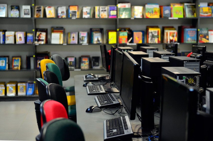 Imagem mostra laboratório de informática com livros em estante ao fundo