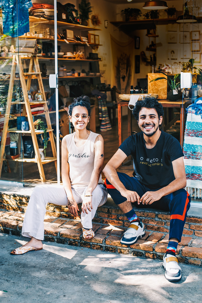 Bia e Bruno sentados no chão da entrada de uma loja da ONG Reviva