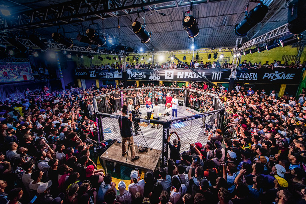 Ringe de MMA com grande público em volta do octógono. No centro, dois rappers com microfones e juizes