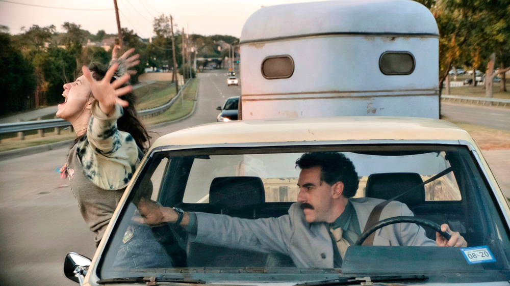 Borat dentro do carro, segurando a filha que está saindo pela janela