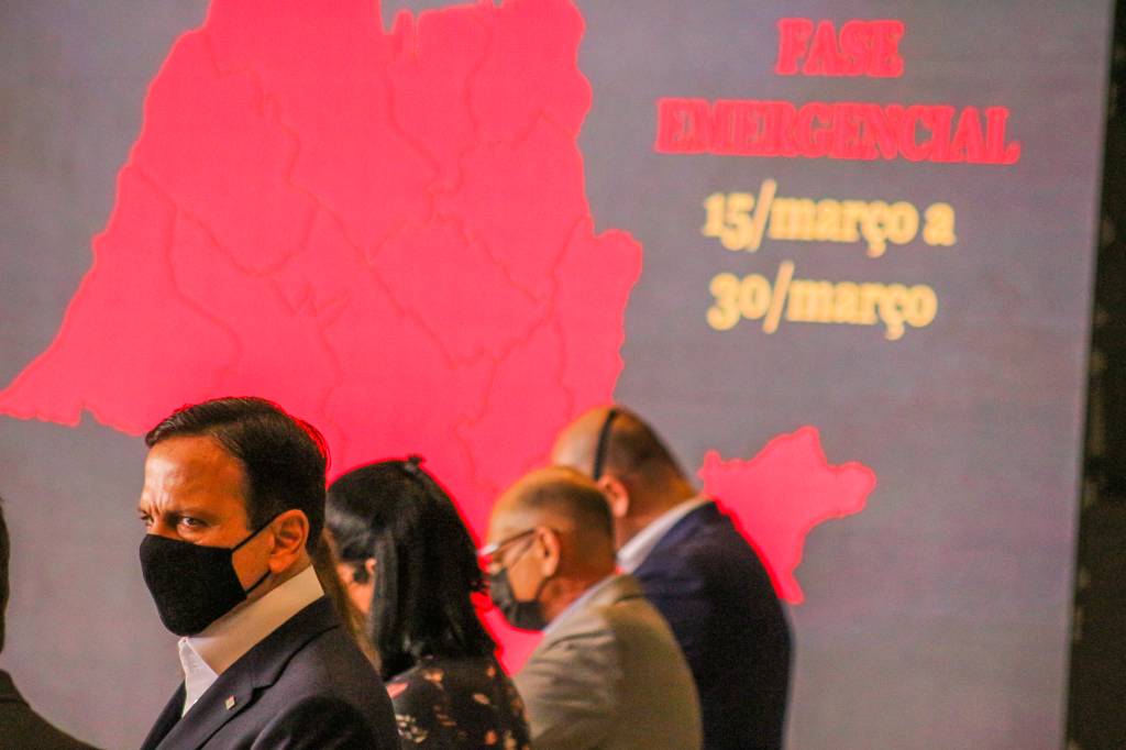 Imagem mostra João Doria em primeiro plano durante coletiva de imprensa; ao fundo, projeção mostra mapa do estado de São Paulo em cor vermelha com frase "fase de restrição" escrita ao lado
