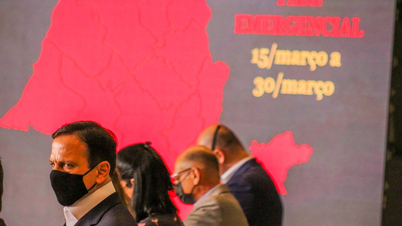 Imagem mostra João Doria em primeiro plano durante coletiva de imprensa; ao fundo, projeção mostra mapa do estado de São Paulo em cor vermelha com frase "fase de restrição" escrita ao lado
