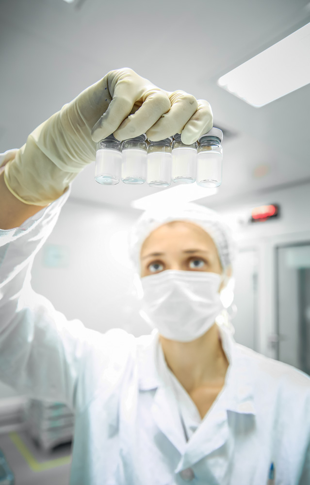 Mulher segura fracos de vacina dentro de laboratório. Ela usa jaleco branco, touca e máscara