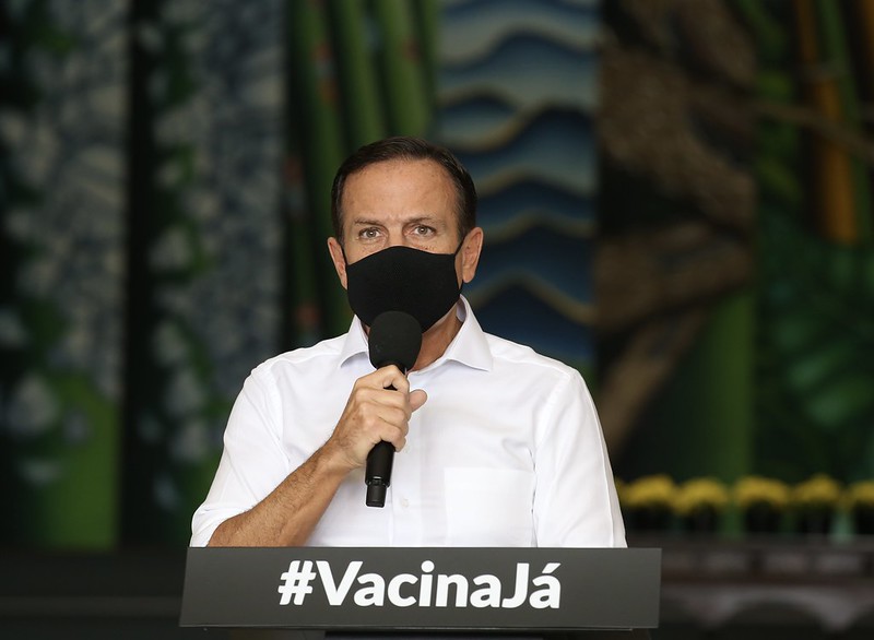 A imagem mostra João Doria, de máscara, segurando um microfone no Palácio dos Bandeirantes. Na mesa em sua frente há a frase "#VacinaJá"