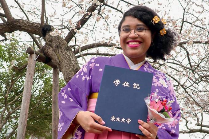 A professora Marina Melo veste quimono roxo e rosa, segura caderno com escritos em japonês e sorri para foto em celebração de formatura de mestrado.
