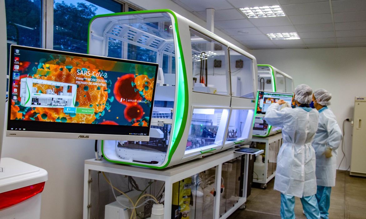 Médicos trabalhando em uma sala branca. Há uma máquina em frente deles, com uma tela ao final dela com imagens do vírus da Covid-19.