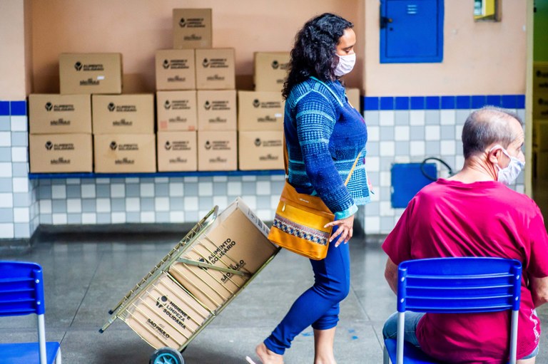 Imagem mostra mulher levando cestas básicas em carrinho de feira