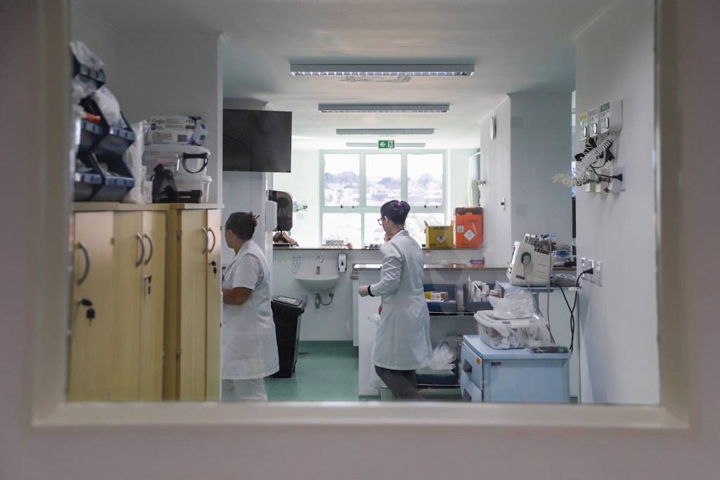 Na imagem é possível ver uma sala de hospital em que há duas mulheres vestindo equipamentos médicos, como jaleco