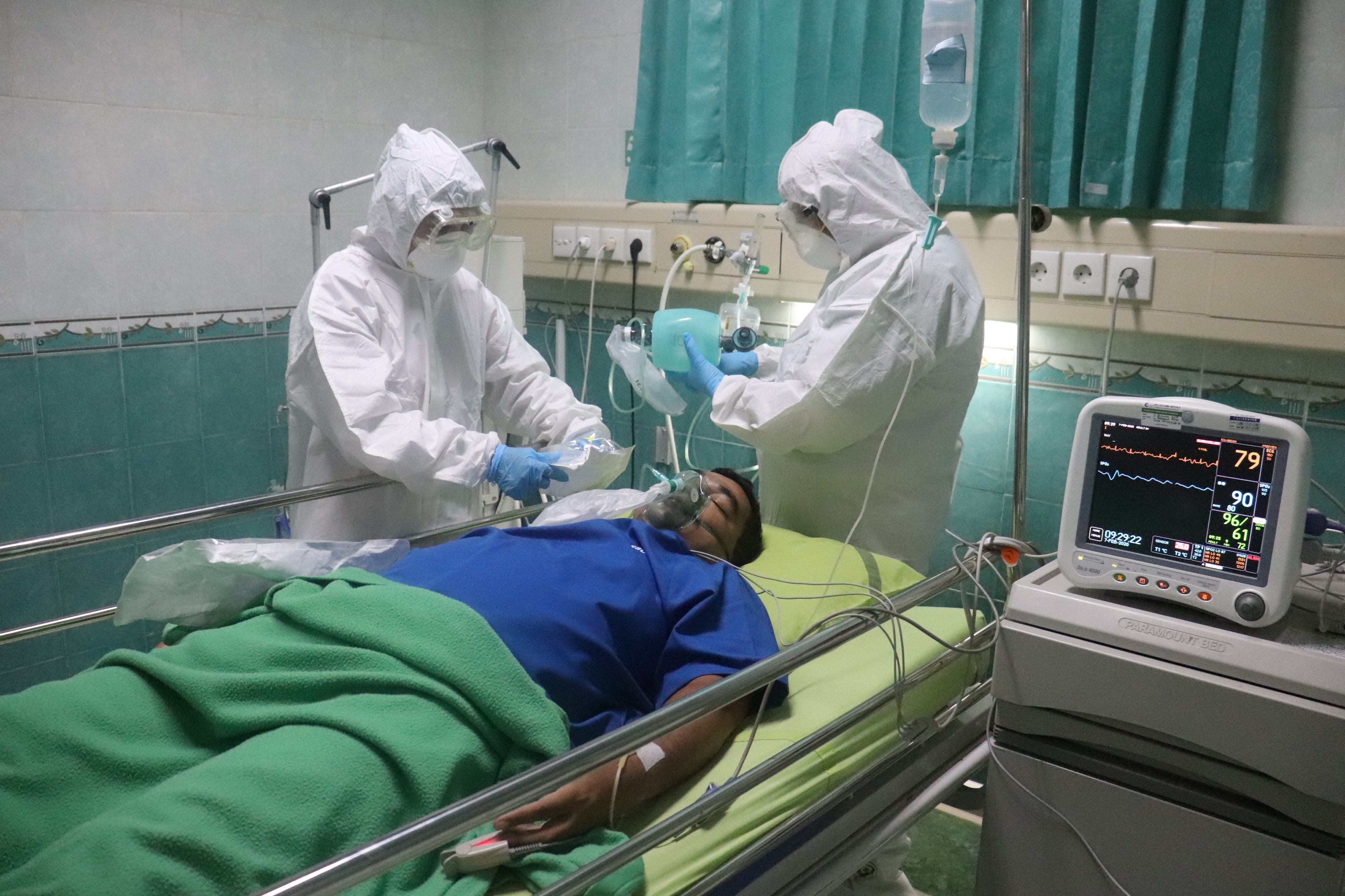 Dois enfermeiros completamente cobertos por roupas brancas de proteção atendem paciente deitado com roupa azul.