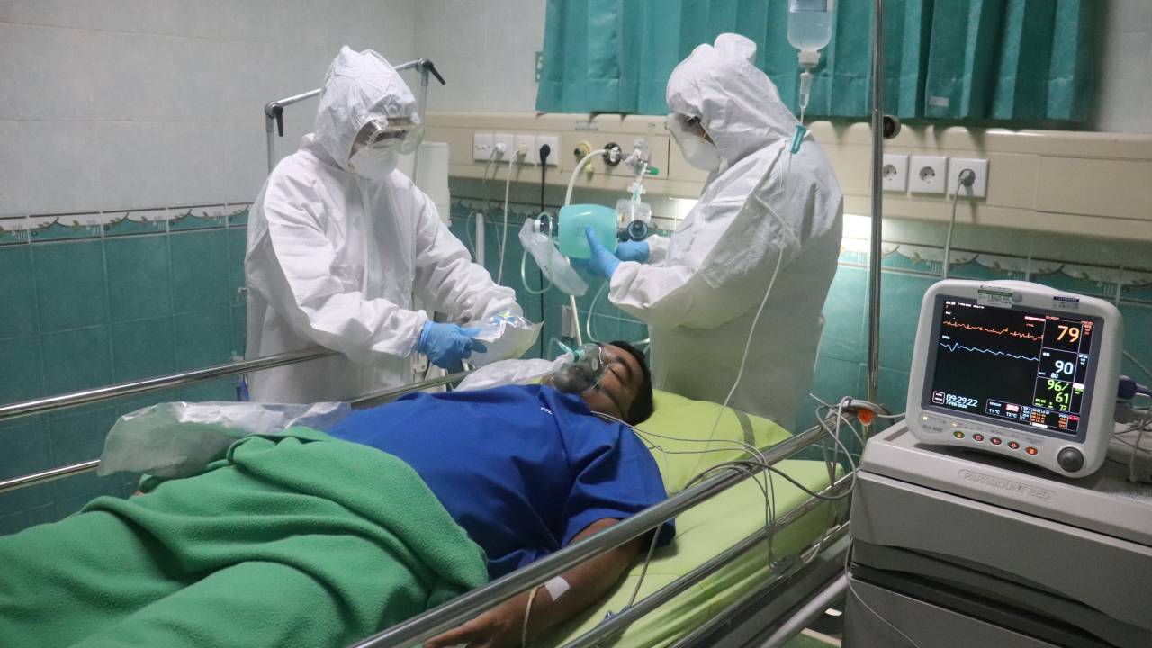 Dois enfermeiros completamente cobertos por roupas brancas de proteção atendem paciente deitado com roupa azul.