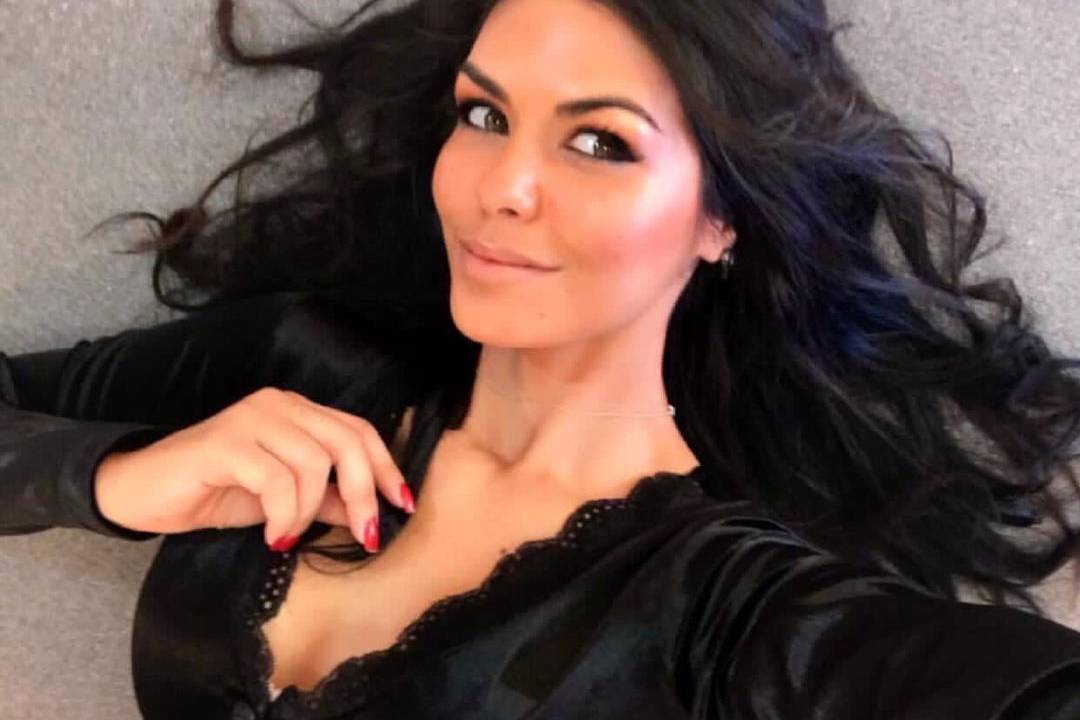 Imagem é uma selfie de Graziela. Ela veste uma blusa preta decotada e cabelos soltos