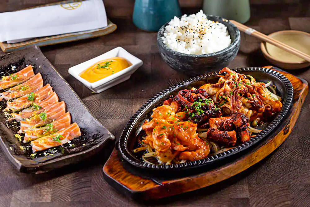 Prato de lula, camarão e polvo marinados em saquê aparecem em destaque na imagem. Ao lado aparece fatias de salmão trufado e arroz branco