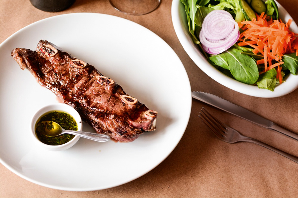 Na imagem aparece uma carne no prato branco, ao lado de um molho. Na parte superior direita da imagem, uma saladinha com folhas, cebola-roxa e cenoura ralada.