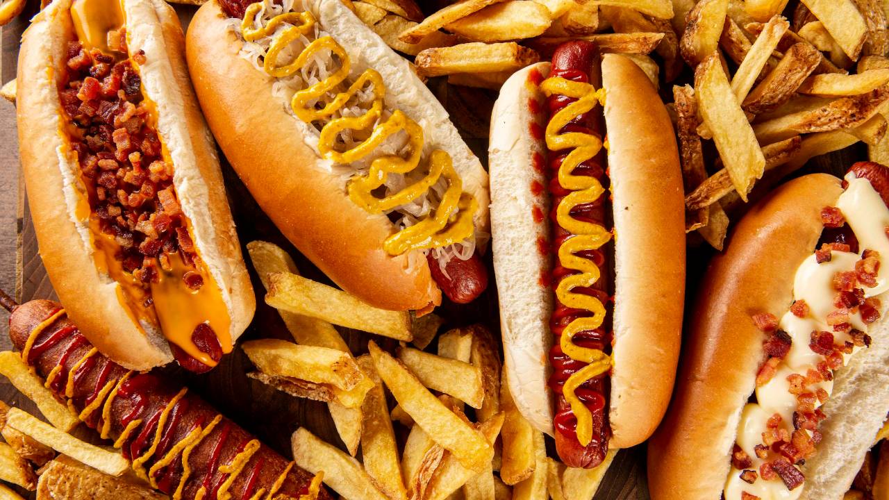 Visão de cima de quatro cachorros-quente sobre mesa rodeados de batata frita e um corndog no canto inferior esquerdo.