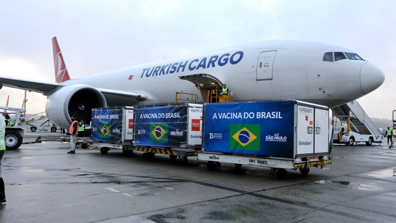 Avião pousado em pista de aeroporto; à frente dele, três caixas grandes que carregam carga de matéria-prima da vacina contra a Covid-19
