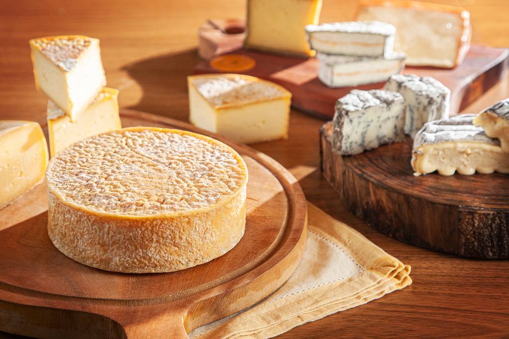 Mesa de madeira com tábuas de queijo. À esquerda em primeiro plano, queijo inteiro redondo sobre tábua do mesmo formato, pedaços em quartos cortados ao fundo. À direita em segundo plano, queijos cobertos por mofo azul cortados ao meio.