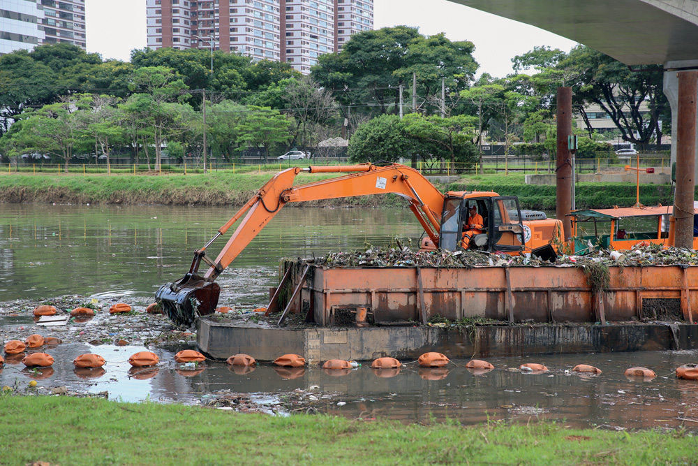Um barco com uma escavadeira na margem do Rio Pinheiro. Há um funcionário dentro, que com o equipamento está removendo objetos de dentro do rio.