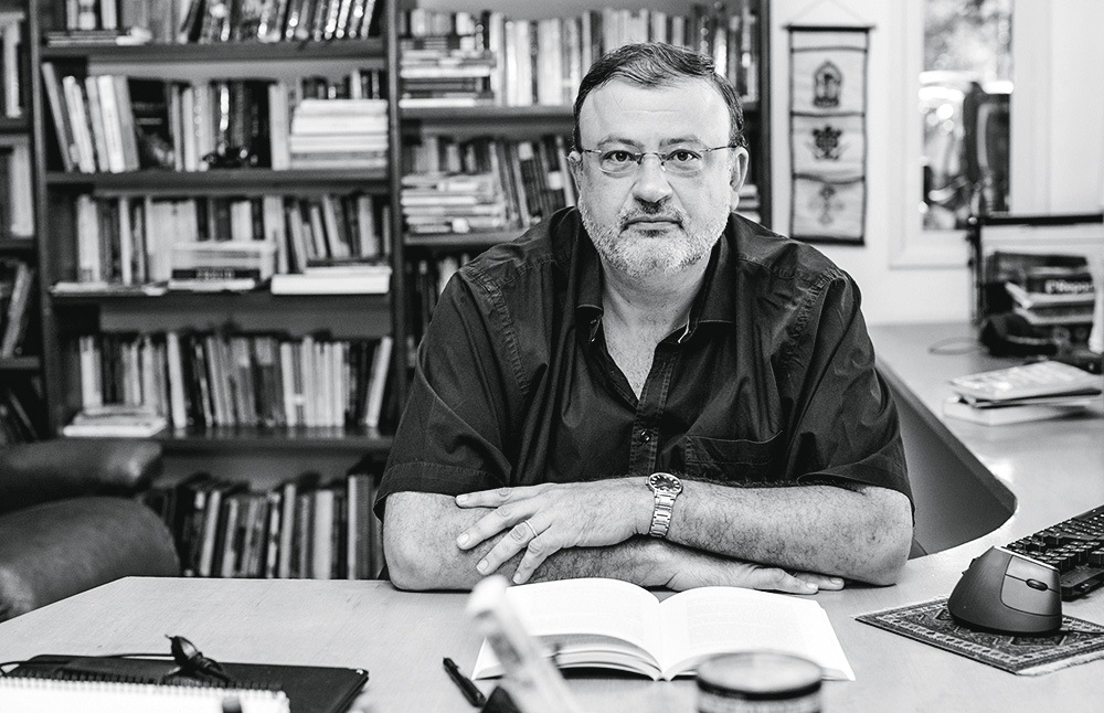 O psicanalista Christian Dunker posa em frente a uma estante de livros sentado em uma cadeira, com mesa na sua frente, que tem um livro aberto. Foto em preto e branco